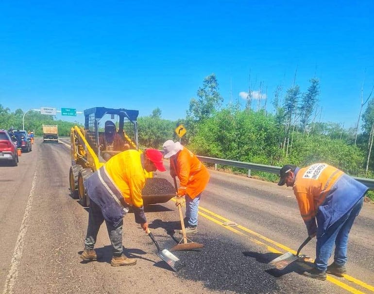 Tres trabajadores en chalecos de seguridad naranjas, usando palas y rodillo en carretera, con vehículos detenidos detrás.