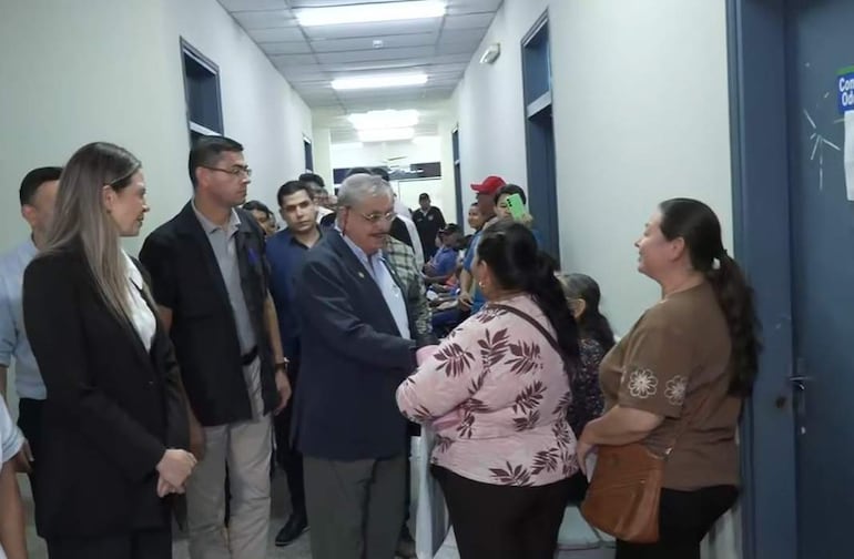El doctor Isaías Fretes, con chaqueta, conversa con una mujer de blusa rosa en un pasillo iluminado, rodeados de personas en espera.