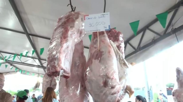 Puesto de venta de carne de cerdo con trozos colgados y un cartel de precio visible, bajo un toldo decorado.