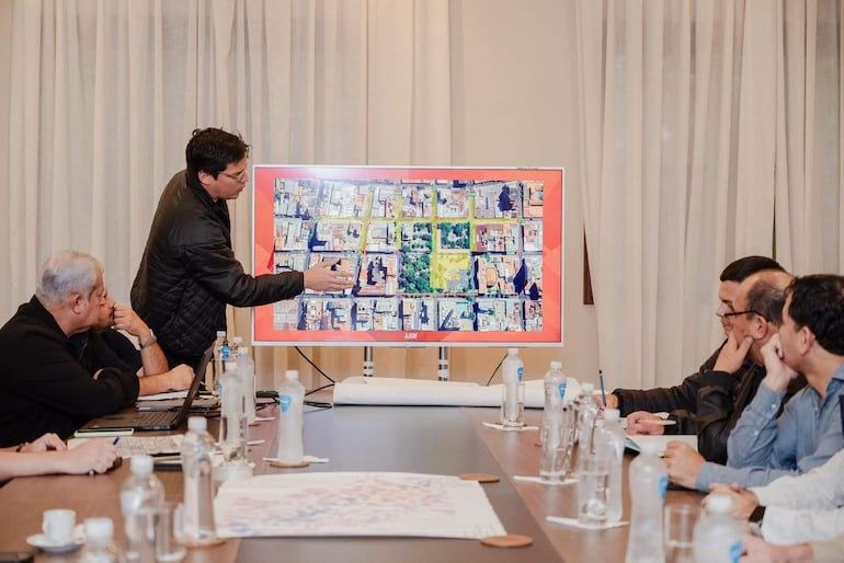 Hombre gesticulando frente a pantalla con mapa, rodeado de cinco hombres atentos, en un entorno iluminado y formal.