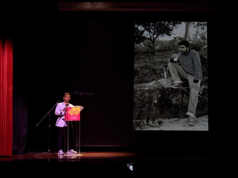 Hombre de pie en podio con chaqueta clara, hablando al público mientras señala una proyección de una fotografía en blanco y negro.
