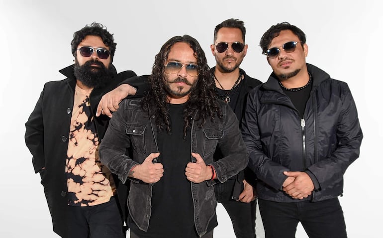 Cuatro hombres de pie en un fondo claro, con chaquetas y gafas de sol, posando con expresiones tranquilas y relajadas.