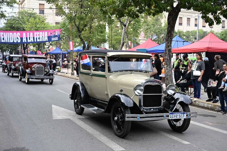 Una mujer sonriente sostiene a un niño en un evento festivo rodeado de autos clásicos y banderas paraguayas.