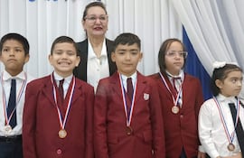 Niños con chaquetas rojas y blancas, sonriendo y serios, en ceremonia con medallas. Mujer detrás con vestido blanco y negro.