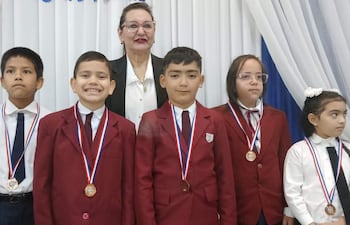 Niños con chaquetas rojas y blancas, sonriendo y serios, en ceremonia con medallas. Mujer detrás con vestido blanco y negro.
