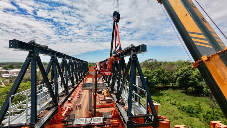 Tres trabajadores en overoles naranjas y cascos colaboran en la construcción, con grúa y estructura metálica en primer plano.