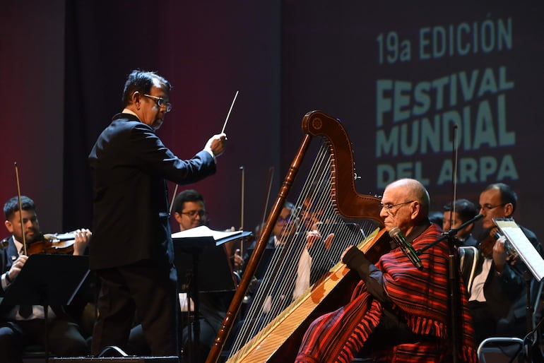 Director de orquesta en saco negro dirige a un arpista vestido de rojo y negro, con músicos de fondo en ambiente formal.