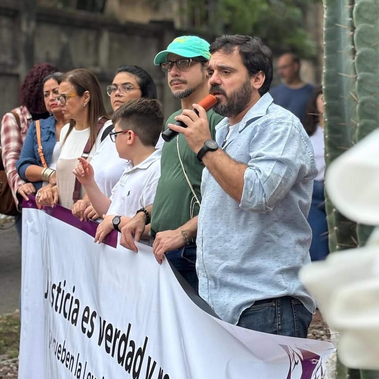 Hombre en gorra verde habla con micrófono, rodeado de adultos y un niño, ante banner de reclamo de justicia.