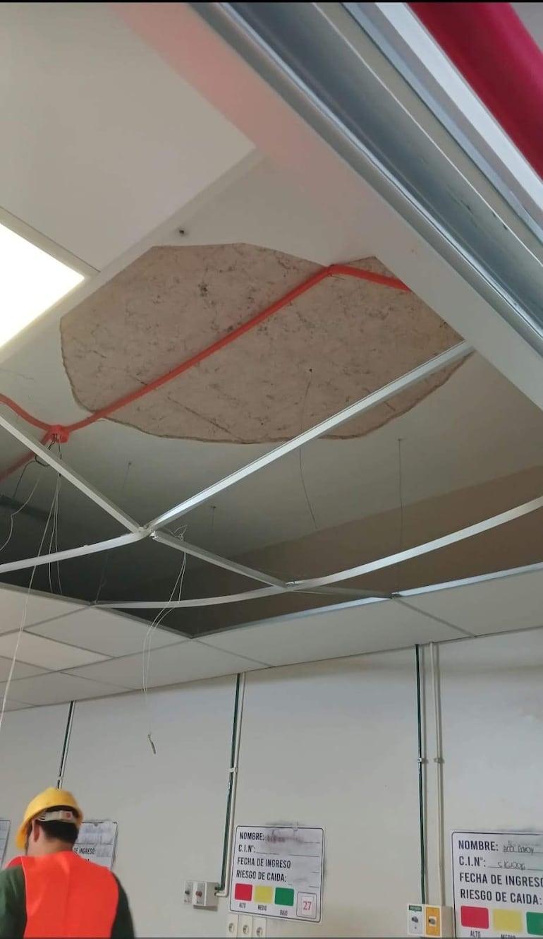 Trabajador con casco amarillo y chaleco naranja en interior dañado, con cables expuestos y carteles de riesgo de caída visibles.