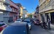 Hombre en motocicleta dialoga con otro en la acera, rodeados de vehículos y edificios coloridos en un día despejado.
