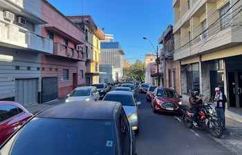 Hombre en motocicleta dialoga con otro en la acera, rodeados de vehículos y edificios coloridos en un día despejado.