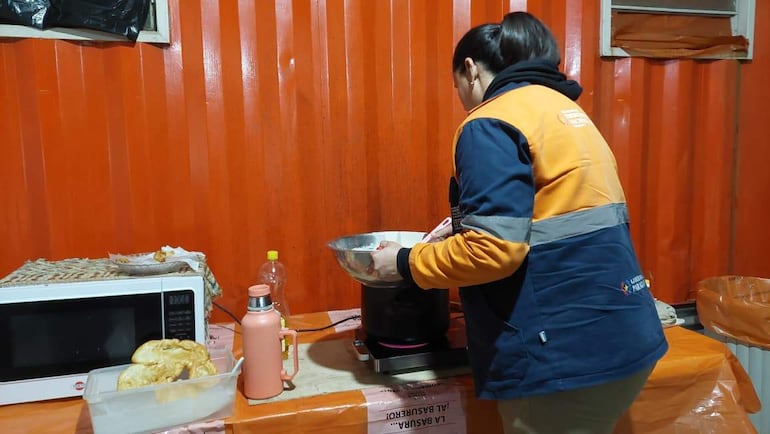 Mujer de espaldas, con chaqueta azul, preparando comida en una cocina con microondas y termos.