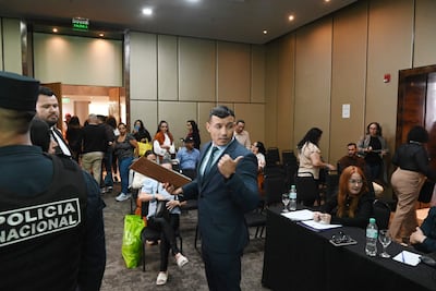Hombre de traje y corbata explica en un evento, con un oficial de policía y público atento en el fondo.