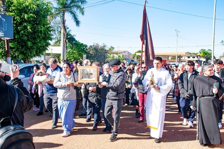 Sacerdote en túnica blanca porta figura religiosa, mientras la multitud viste abrigos y algunos graban el evento en la calle.
