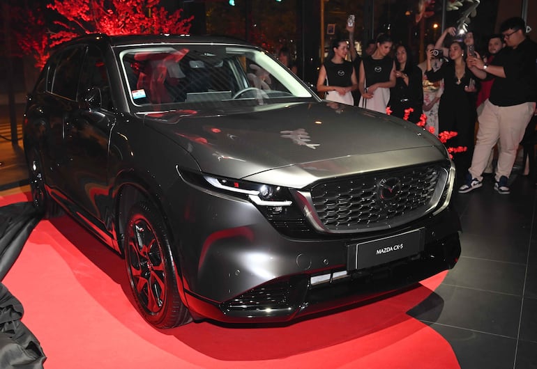 Un automóvil gris Mazda CX-5 se exhibe en un showroom decorado con luces rojas, mientras asistentes observan y toman fotos.