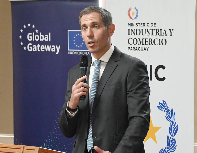 Hombre con traje oscuro y corbata clara habla con micrófono en mano, banners del Ministerio de Industria y Comercio y Unión Europea en el fondo.