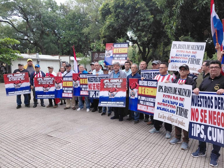 Hombres con pancartas como 'IPS, CUMPLAI' y 'NO SE NEGOCIAN' en la plaza, rodeados de árboles y edificios.