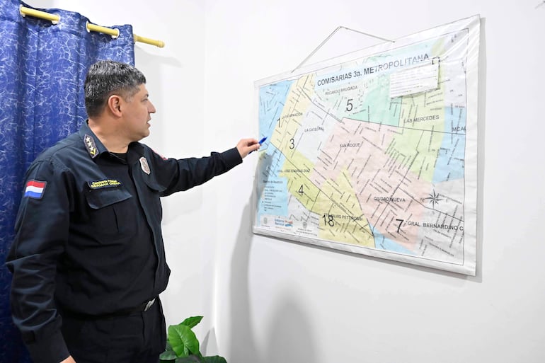 Oficial de policía en uniforme revisa un mapa en una oficina con cortina azul y planta verde.
