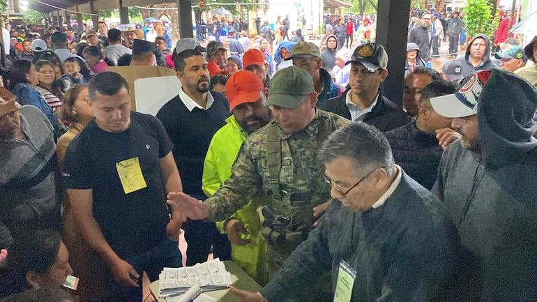 Hombre de chaqueta clara discute con grupo, mientras observan autoridades en evento al aire libre bajo techo.