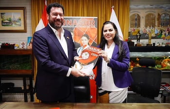Cappello, con chaqueta oscura y barba, sonríe mientras entrega un documento a Reyes, quien viste un saco morado.