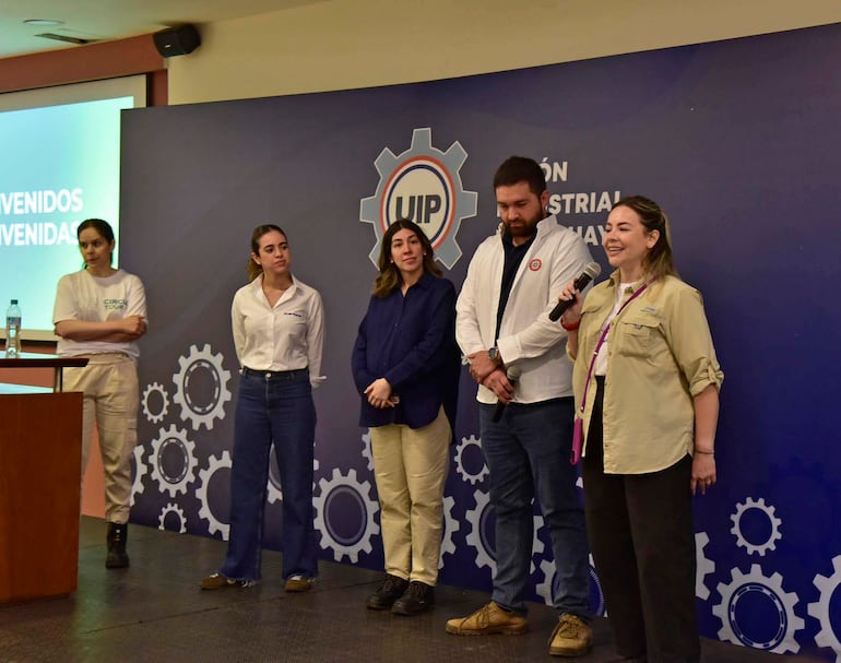 Cinco personas en un evento: mujeres con camisetas claras y hombre con chaqueta blanca, orando y escuchando en fondo gráfico industrial.
