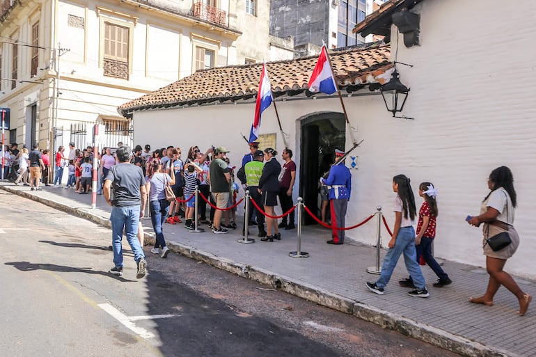 Dos guardias en uniformes ceremoniales sostienen banderas paraguayas, mientras personas de diversos colores esperan en fila bajo el sol.