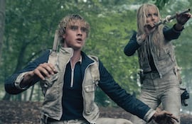 Actores en abrigo y chaquetas, con expresiones de sorpresa y miedo, rodeados de un ambiente de acción con armas visibles.