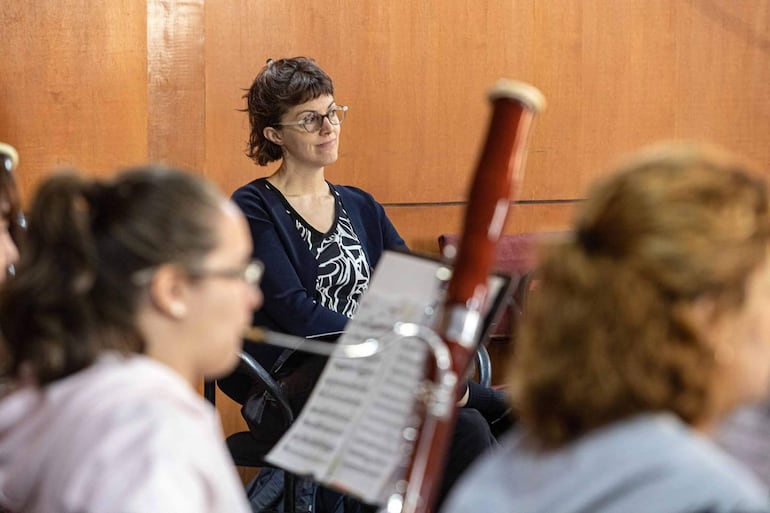 Mujer de cabello corto y ondulado, con gafas, escucha atentamente mientras otros sostienen instrumentos musicales en un aula.
