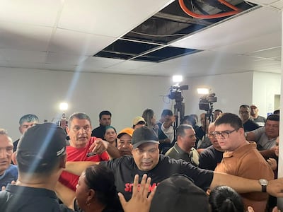 Juan Carlos Meza con camiseta negra y gesticulando mientras otros escuchan atentamente en un ambiente tenso y concurrido.