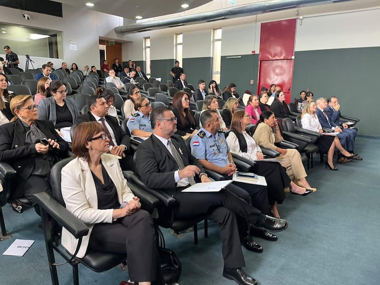 Mujer con gafas y chaqueta blanca en primer plano, rodeada de personas escuchando atentamente en sala de conferencias.