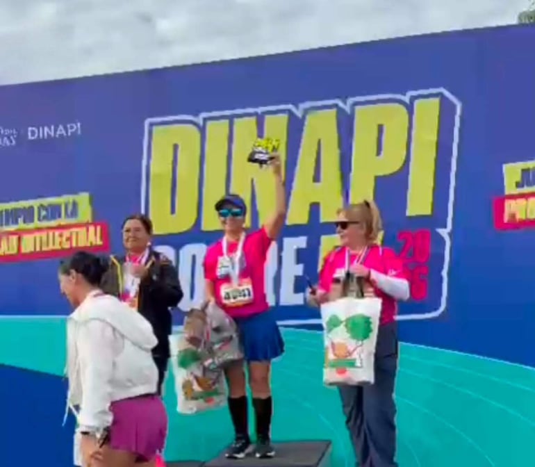 Mujeres sonrientes en la premiación, una con trofeo y otra con medalla, en un ambiente festivo al aire libre.