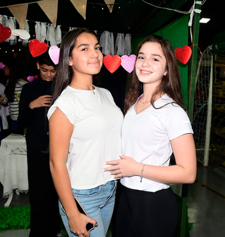Dos mujeres sonrientes con camisetas blancas, rodeadas de decoraciones de corazones, mientras un joven observa al fondo.