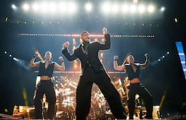 Ricky Martin en atuendo oscuro sonríe y canta, acompañado de dos bailarines en ropa sin mangas, bajo luces brillantes.