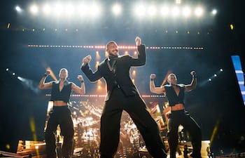 Ricky Martin en atuendo oscuro sonríe y canta, acompañado de dos bailarines en ropa sin mangas, bajo luces brillantes.