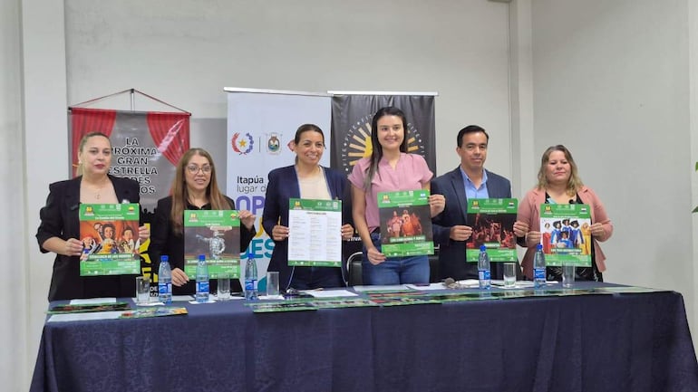 Seis personas de pie detrás de una mesa, sosteniendo carteles verdes. Tres mujeres y tres hombres, ambiente festivo y cultural.