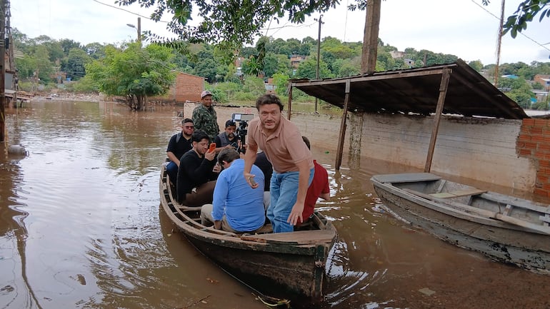 Seis personas en canoa, algunos con teléfonos móviles, en un entorno inundado, con agua estancada y viviendas sumergidas.