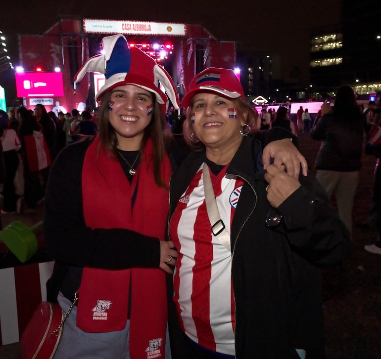 Dos mujeres sonríen, una con gorro y bufanda roja, la otra con camiseta de selección paraguaya, en un ambiente festivo.
