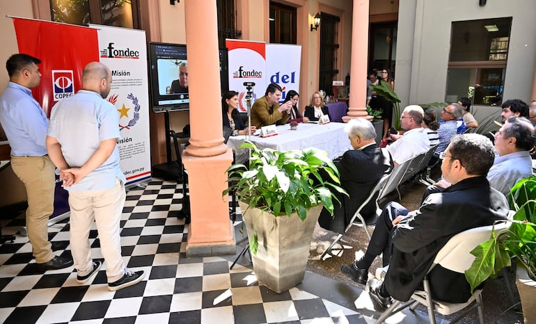 Cinco personas sentadas en mesa blanca, hablando y escuchando atentamente, decorado con plantas y pantallas en un ambiente relajado.