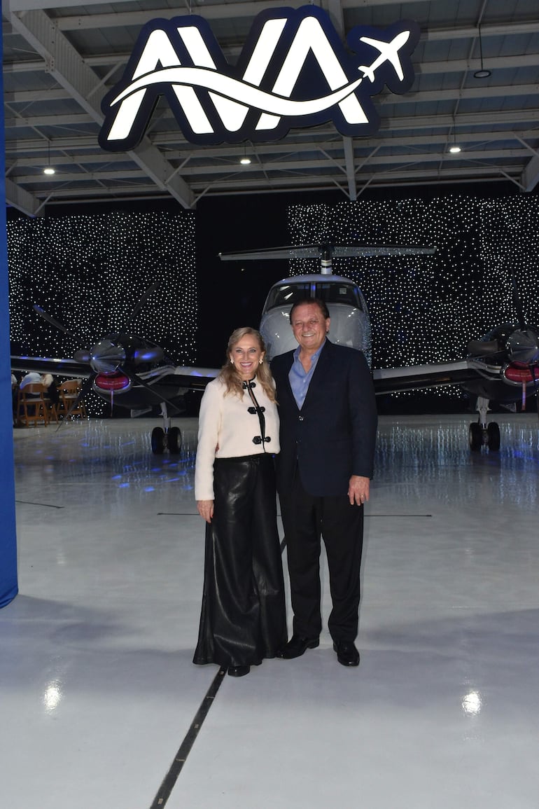 Andrés Trociuk y una mujer sonriente, bien vestidos, posan en un hangar con un avión y luces brillantes de fondo.