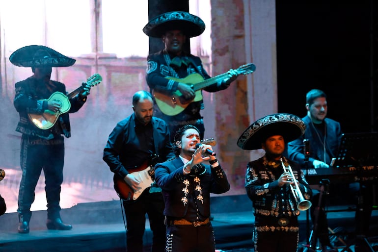Alex Fernández, vestido de mariachi, canta con emoción mientras guitarristas y trompetistas lo acompañan en un escenario iluminado.