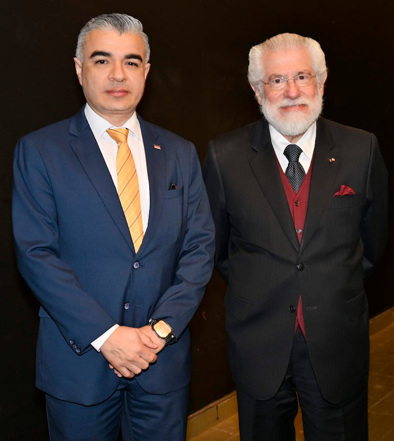 Hombre con traje azul y corbata amarilla junto a otro con traje negro y chaleco rojo, ambos en un ambiente formal.