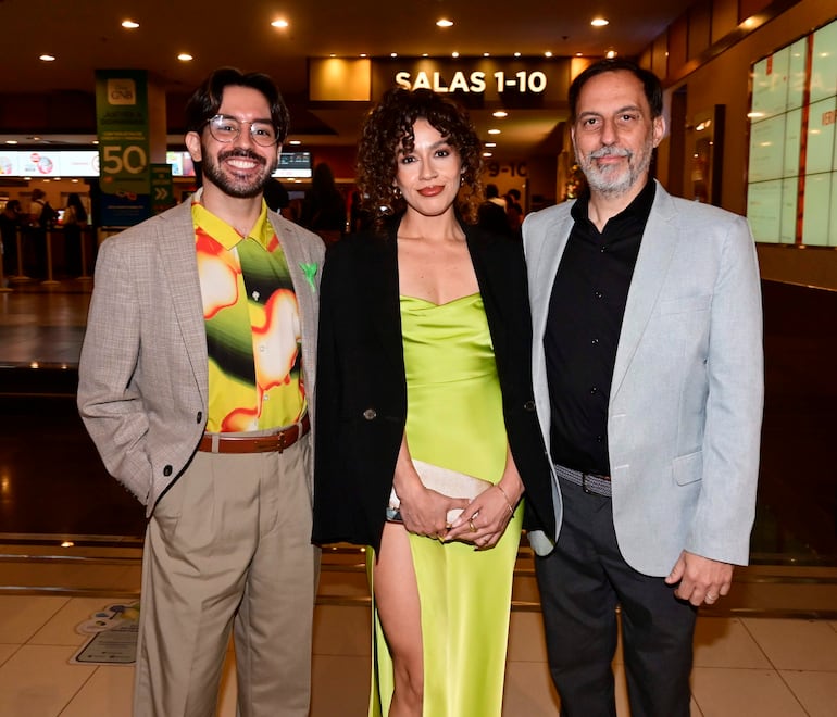 Una mujer en vestido verde luminoso y abrigo negro flanqueada por dos hombres en trajes, posando en un cine.