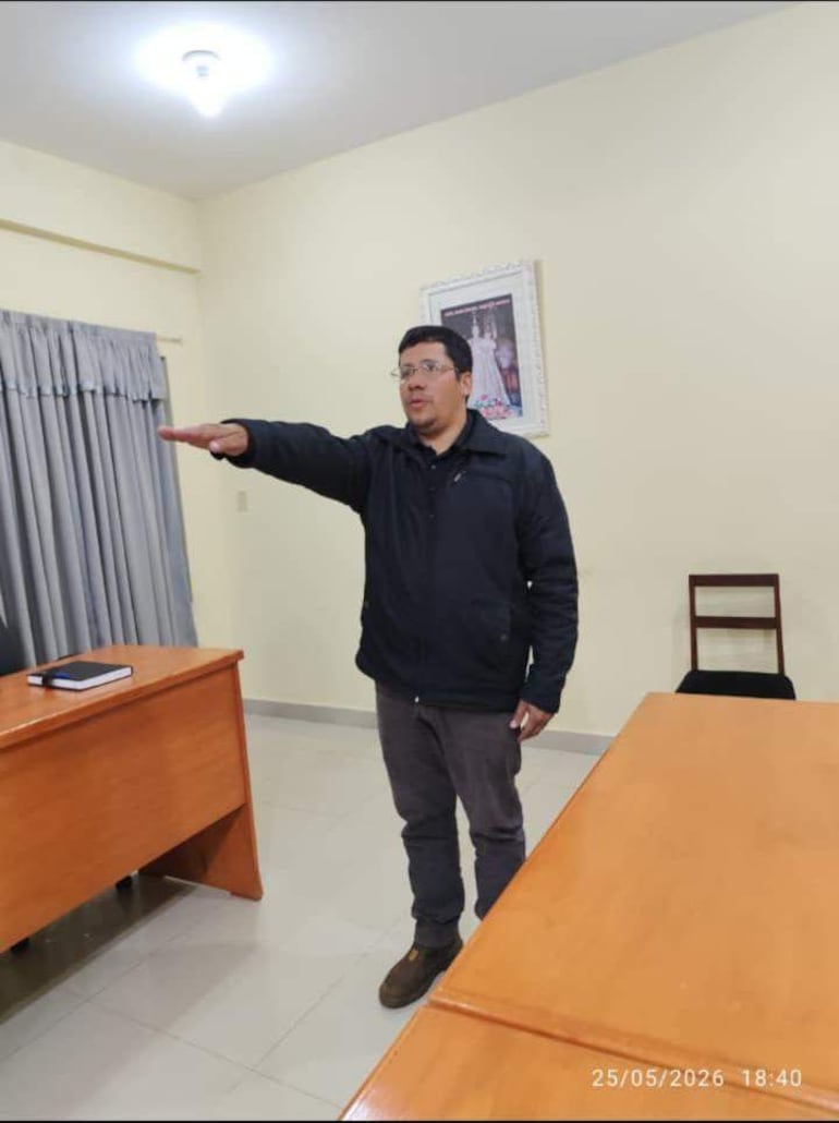Hombre de pie en sala de reuniones, vistiendo abrigo negro y señalando con la mano. Fondo con cortina gris y cuadro en la pared.