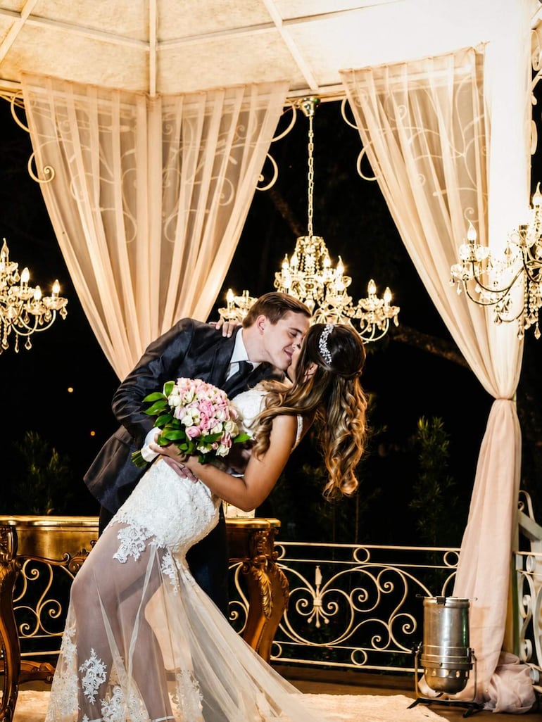 Pareja de recién casados, hombre en traje negro y mujer en vestido blanco con ramo rosa, celebran con alegría en elegante entorno con candelabros.
