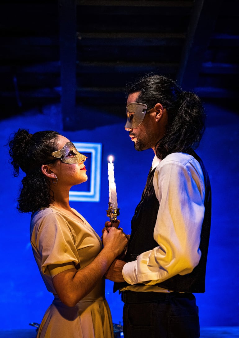 Actores con máscaras, uno en blusa clara y chaleco oscuro, sostienen una vela en un ambiente teatral azul profundo.