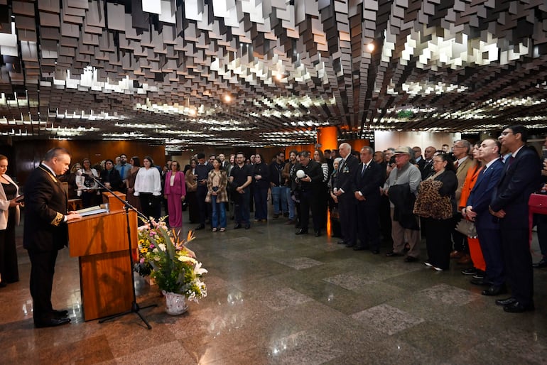 Hombre en uniforme en podio con flores, mientras el público escucha atentamente en un ambiente elegante.