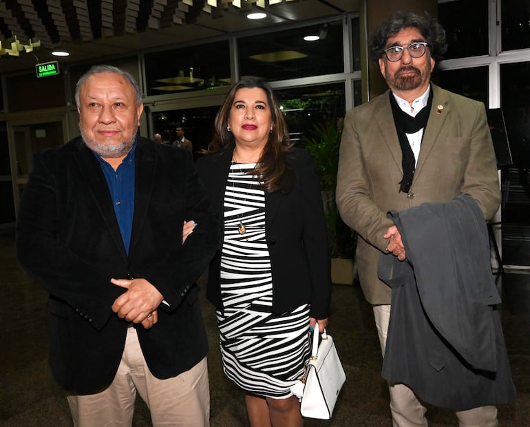 Hombre canoso con blazer negro, mujer en vestido blanco y negro, hombre con abrigo gris sosteniendo chaqueta, en ambiente elegante.