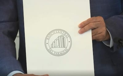 Mano sosteniendo documento del Banco Central del Paraguay, con logotipo impreso en círculo sobre fondo blanco.