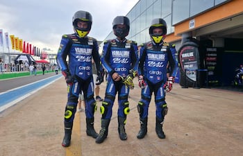 Tres motociclistas en trajes azules de Yamaha posan con confianza en un circuito, rodeados de ambiente animado y banderas.