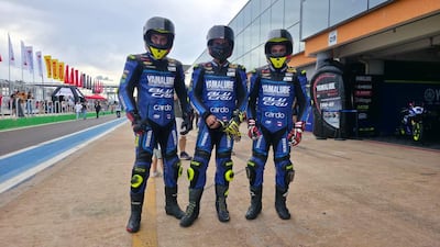 Tres motociclistas en trajes azules de Yamaha posan con confianza en un circuito, rodeados de ambiente animado y banderas.